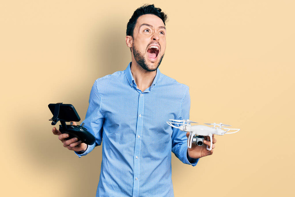 Hombre hispano con barba usando dron con control remoto enojado y loco gritando frustrado y furioso, gritando con ira mirando hacia arriba.  - Foto, Imagen