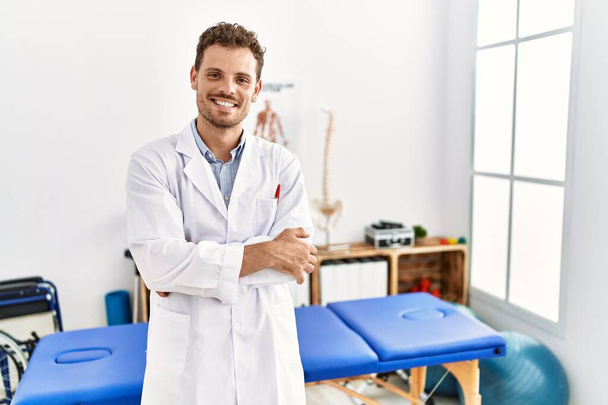Przystojny młodzieniec pracujący w klinice uzdrawiania bólu uśmiechnięty ze skrzyżowanymi rękami patrzący w kamerę. osoba pozytywna.  - Zdjęcie, obraz