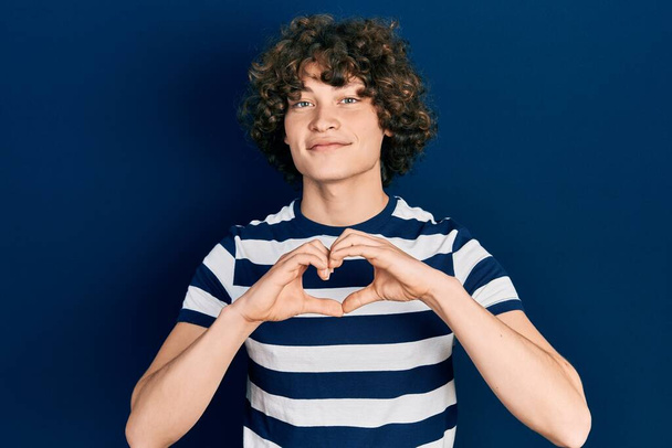 Красивый молодой человек в обычной полосатой футболке, улыбающийся в любви, делает символическую форму сердца руками. романтическая концепция.  - Фото, изображение