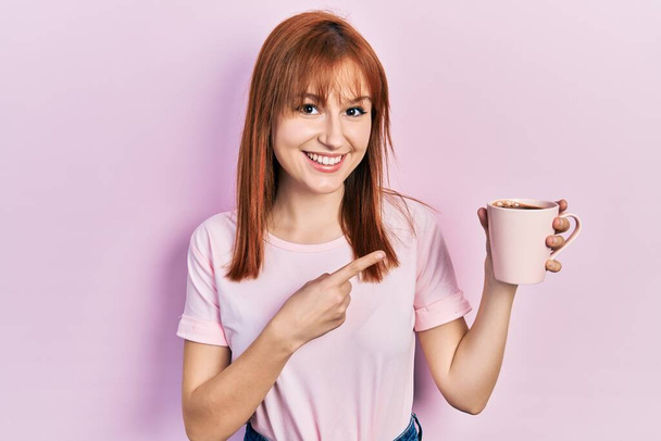 Рыжая молодая женщина пьет чашку кофе улыбаясь счастливо указывая рукой и пальцем  - Фото, изображение