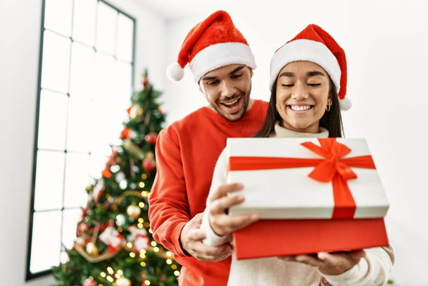 Młoda Latynoska para uśmiechnięta szczęśliwa i przytulająca się do świątecznego prezentu w domu. - Zdjęcie, obraz