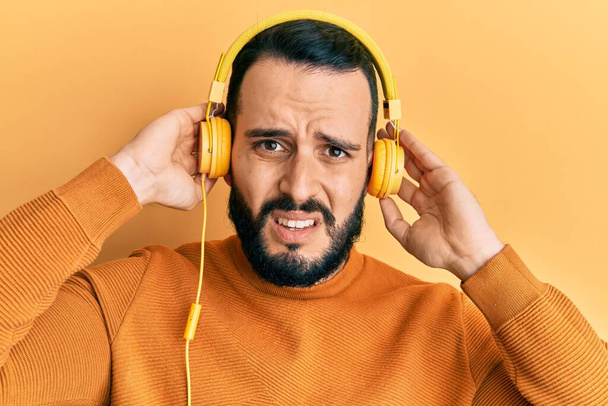 Joven con barba escuchando música usando auriculares sin pistas y expresión confusa. concepto de duda.  - Foto, imagen
