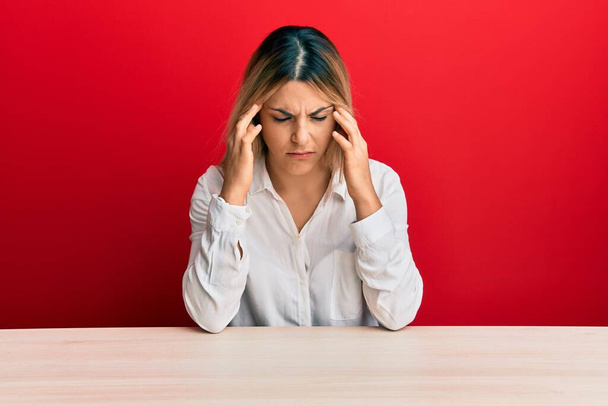 Junge kaukasische Frau in lässiger Kleidung sitzt auf dem Tisch mit der Hand auf dem Kopf vor Schmerzen im Kopf wegen Stress. Migräne.  - Foto, Bild