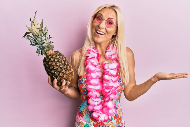 Młoda blondynka ubrana w strój kąpielowy i hawajskie lei trzymająca ananasa świętującego osiągnięcie szczęśliwym uśmiechem i wyrazem zwycięzcy z podniesionymi dłońmi  - Zdjęcie, obraz