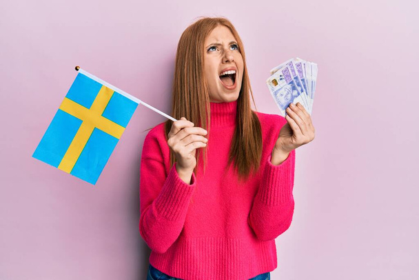 Mladá Irka držící švédskou vlajku a korunové bankovky rozzlobená a šíleně křičící, frustrovaná a rozzuřená, křičící vzteky vzhlížející.  - Fotografie, Obrázek