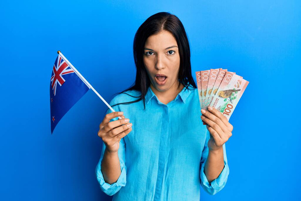 Giovane donna latina in possesso di bandiera neozelandese e dollari in faccia shock, guardando scettico e sarcastico, sorpreso a bocca aperta  - Foto, immagini