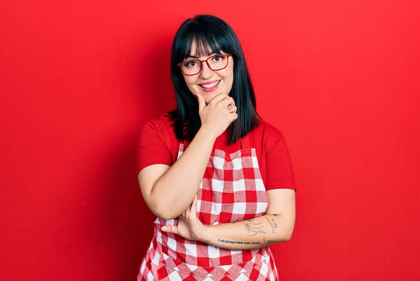 Young hispanic woman wearing cook apron and glasses looking confident at the camera smiling with crossed arms and hand raised on chin. thinking positive.  - Photo, Image