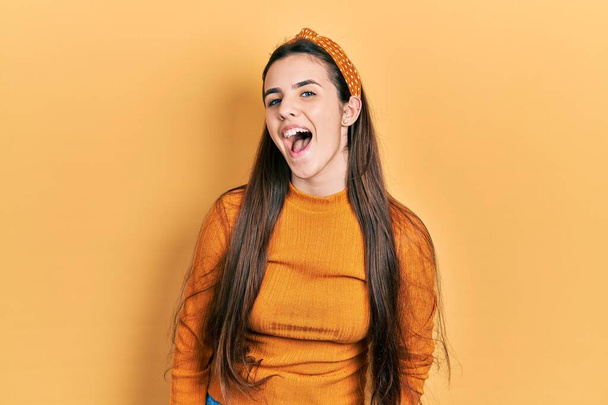 Joven adolescente morena vistiendo suéter amarillo casual sonriendo y riendo en voz alta porque divertido chiste loco con las manos en el cuerpo.  - Foto, Imagen