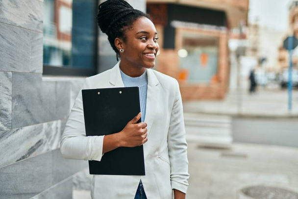 Joven mujer de negocios afroamericana sonriendo feliz sujetando el portapapeles en la ciudad. - Foto, Imagen
