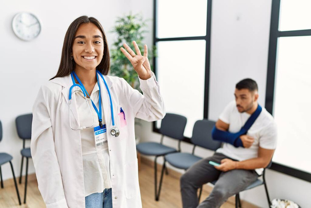 Fiatal ázsiai orvos nő a váróteremben egy törött karú férfival, aki a négyes számú ujjal mutogat, miközben magabiztosan és boldogan mosolyog..  - Fotó, kép