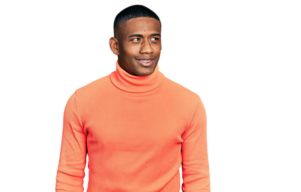 Junger schwarzer Mann in orangefarbenem Rollkragenpullover, der mit einem Lächeln im Gesicht und natürlichem Gesichtsausdruck zur Seite schaut. Lachen selbstbewusst.  - Foto, Bild