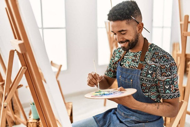 Νεαρός Αφροαμερικάνος καλλιτέχνης χαμογελά χαρούμενος ζωγραφική στο στούντιο τέχνης. - Φωτογραφία, εικόνα