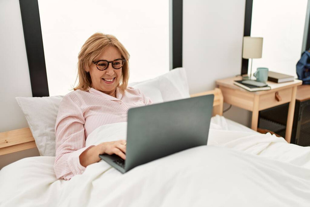 Блондинка середнього віку посміхається щаслива, використовуючи ноутбук, що лежить на ліжку вдома
. - Фото, зображення