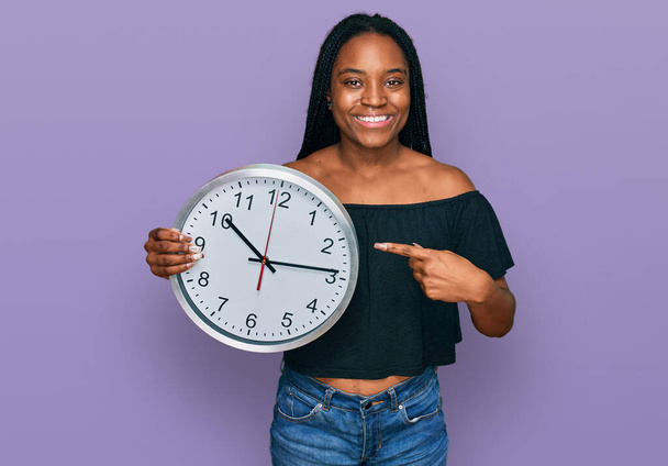 Młoda Afroamerykanka trzyma duży zegar uśmiechając się szczęśliwy wskazując ręką i palcem  - Zdjęcie, obraz