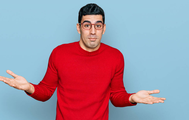 Handsome hispanic man wearing casual clothes and glasses clueless and confused expression with arms and hands raised. doubt concept.  - Photo, Image