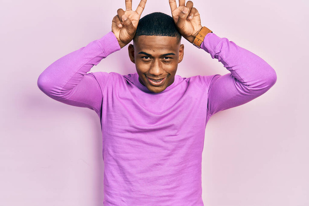 Νέοι μαύροι φορώντας casual ροζ πουλόβερ θέτουν αστείο και τρελό με τα δάχτυλα στο κεφάλι ως αυτιά λαγουδάκι, χαμογελώντας χαρούμενα  - Φωτογραφία, εικόνα