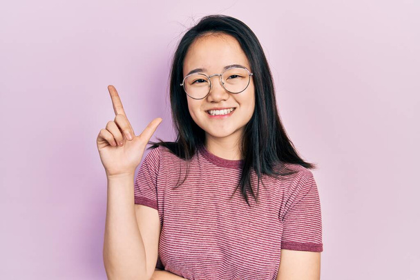 Fiatal kínai lány alkalmi ruhát és szemüveget visel, nagy mosollyal az arcán, mutatva kézzel és ujjal oldalra a kamerába nézve..  - Fotó, kép