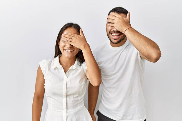 Young interracial couple standing together in love over isolated background smiling and laughing with hand on face covering eyes for surprise. blind concept.  - Photo, Image
