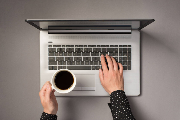 Vista superior en primera persona foto de las manos de la mujer escribiendo en el teclado del ordenador portátil y sosteniendo la taza de café sobre un fondo gris aislado - Foto, Imagen