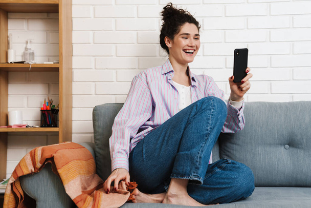 Happy mid aged brunette woman holding mobile phone while sitting on a couch at home, on a video call - Foto, Bild
