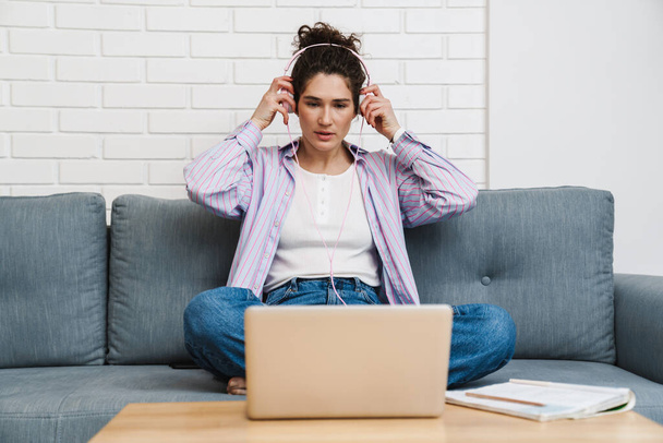The curly-haired woman putting on headphones and looking into a laptop monitor while sitting at a table on a sofa in a bright room - Photo, Image