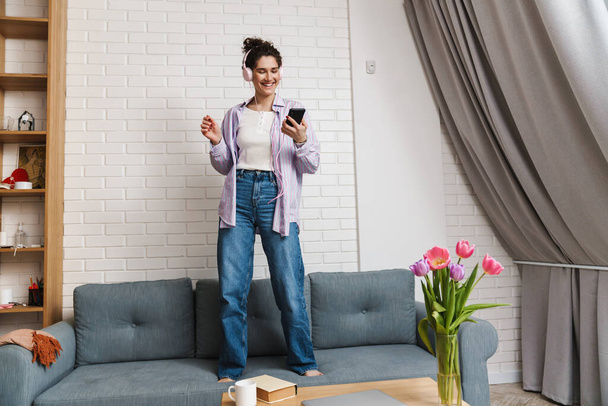 The smiling woman listening to music in headphones while standing on sofa and holding phone in hand in the room - Foto, imagen