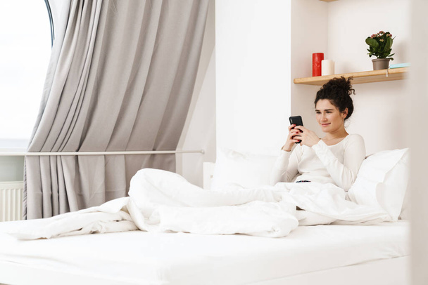 Smiling mid aged brunette woman on a video call via mobile phone while laying in bed - Fotó, kép