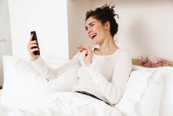 Smiling mid aged brunette woman on a video call via mobile phone while laying in bed - Photo, image