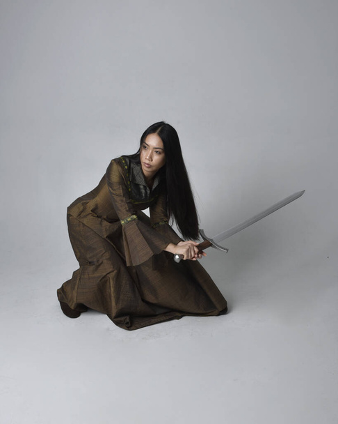 Full length portret van mooie jonge Aziatische vrouw met lang haar in middeleeuwse fantasie toga kostuum. Genadige zittende houding met een zwaard, geïsoleerd op studio-achtergrond. - Foto, afbeelding