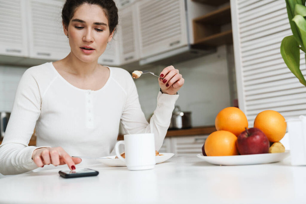 A pensive woman holding a cut piece of something on a fork and typing at the phone while sitting at a table in a bright kitchen - Photo, image