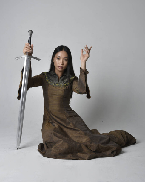 Ganzes Porträt einer schönen jungen asiatischen Frau mit langen Haaren, die ein mittelalterliches Fantasiegewand trägt. Anmutige Sitzhaltung mit einem Schwert, isoliert auf Studiohintergrund. - Foto, Bild