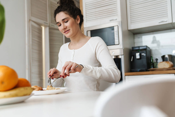 Усміхнена жінка ріже їжу на тарілці, сидячи за столом на яскравій кухні
 - Фото, зображення