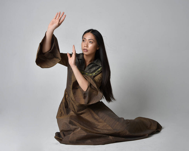 Full length portret van mooie jonge Aziatische vrouw met lang haar in middeleeuwse fantasie toga kostuum. Gracieuze zithouding, geïsoleerd op studioachtergrond. - Foto, afbeelding