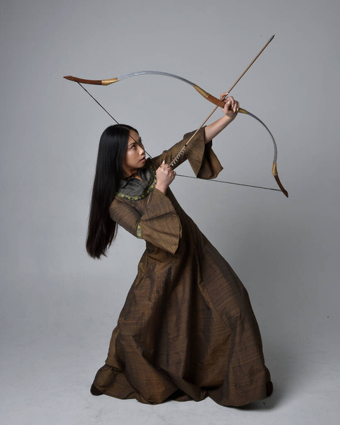 Close up  portrait of beautiful young asian woman with long hair wearing medieval fantasy gown. Graceful pose holding a long bow and arrow,  isolated on studio background. - Фото, зображення