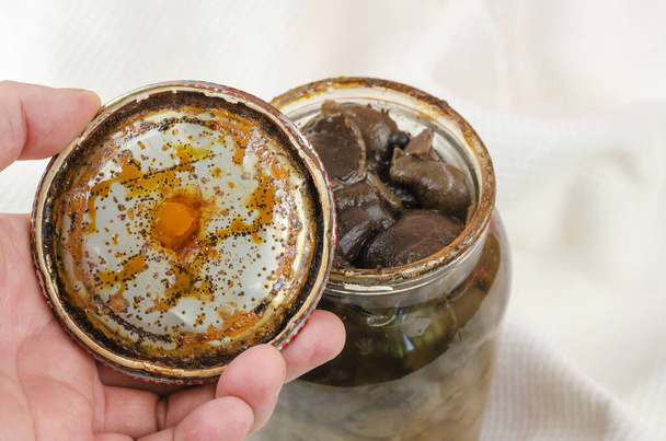 Botulism, food poisoning from canned foods concept. Handholds Rusty tin lid with black stains. Opened Glass jar with homemade pickled Suillus. Spoiled blackened forest mushrooms - Photo, Image
