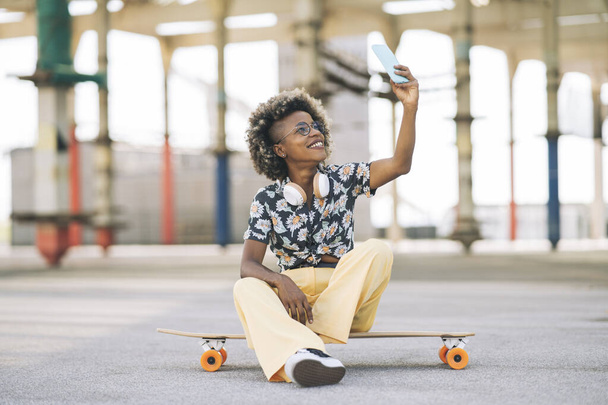pretty blonde afro woman in glasses with skateboard, sitting takes a photo with smartphone - Photo, Image