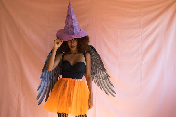 junge afrikanisch-amerikanische Frau im Hexenkostüm zur Halloween-Party. Sie trägt einen Hexenhut, orangefarbenen Rock und schwarze Engelsflügel. Die Frau lächelt in die Kamera, während sie in verschiedenen Posen posiert. - Foto, Bild