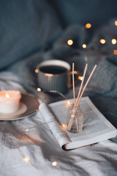 Bottle with liquid home fragrance and wooden bamboo sticks, scentes burn candle and cup of black tea over glow lights at background close up. Winter holiday season. Cozy romantic home atmosphere.  - Photo, Image