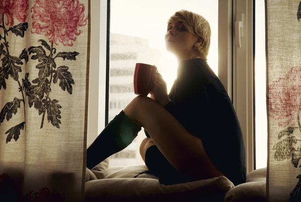 Portrait of charming girl sitting on windowsill and looking through window in the dusk - Foto, afbeelding