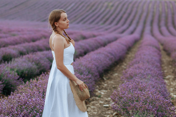 A beautiful young woman in a white dress with a hat walks through a lavender field - Photo, Image