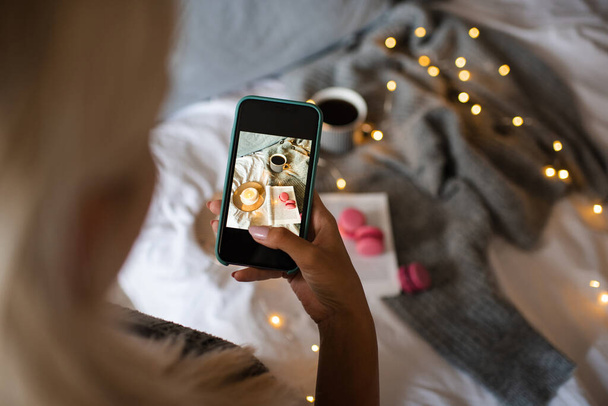 Frau macht Bild auf Smartphone von Winter gemütliche Wohnung Interieur über Weihnachtsbeleuchtung aus nächster Nähe. Herbstzeit. - Foto, Bild