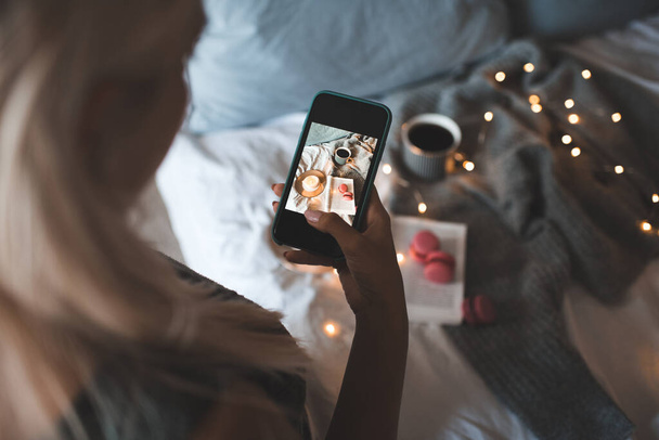 Frau macht Bild auf Smartphone von Winter gemütliche Wohnung Interieur über Weihnachtsbeleuchtung aus nächster Nähe. Herbstzeit. - Foto, Bild