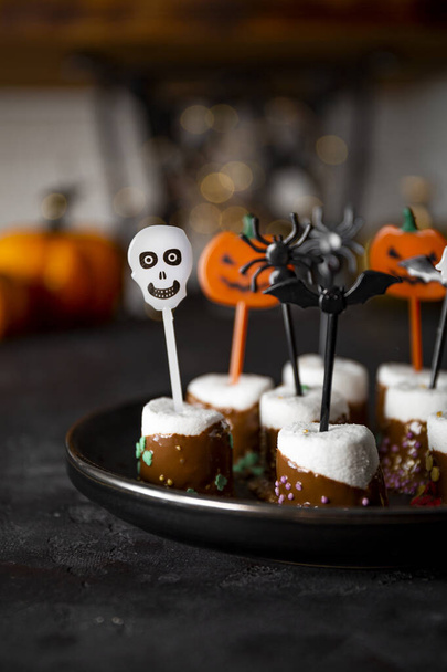 Sweet Halloween treat, chocolate covered marshmallows with sugar and Halloween decorations on a black plate, vertical photo with soft focus - Photo, image