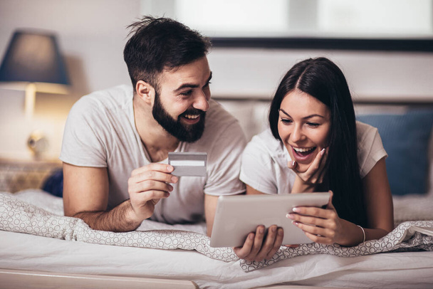 素敵な若い幸せなカップルがベッドに横たわって、クレジットカードとデジタルタブレットでオンラインショッピング. - 写真・画像