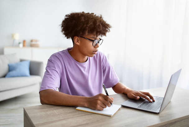 Conceito de e-learning. Adolescente afro-americano inteligente escrevendo em notebook durante lição na web em casa - Foto, Imagem