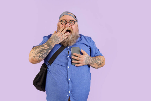 Surprised obese man holds mobile phone covering mouth by palm on purple background - Fotografie, Obrázek