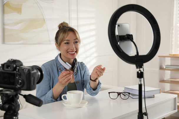Blogger with cup of tea recording video at table indoors. Using ring lamp and camera - 写真・画像