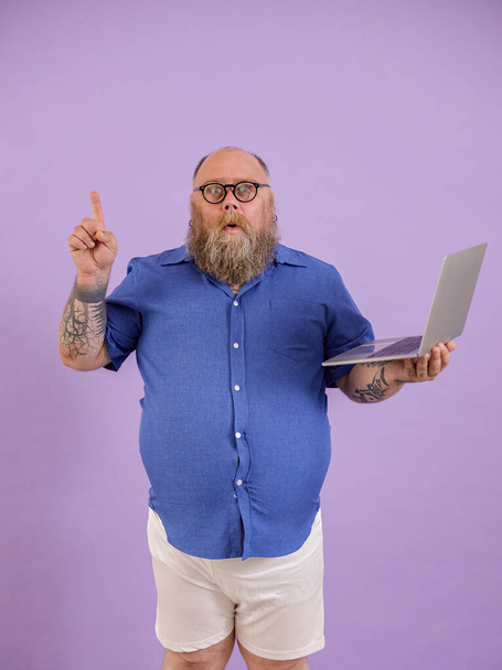 Hombre divertido con sobrepeso consiguió idea interesante sosteniendo el ordenador portátil sobre fondo púrpura - Foto, imagen