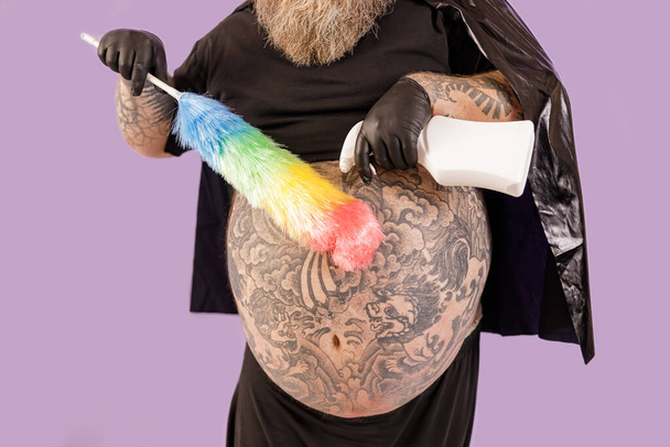 Grubas w garniturze bohatera spryskuje detergentem szczotkę na fioletowym tle - Zdjęcie, obraz