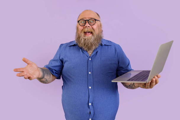 Joyful fat businessman in tight shirt with spectacles holds laptop on purple background - Photo, Image